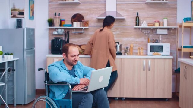 Disabled man waving on video conference using laptop in kitchen. Corporate man with paralysis handicap disability handicapped difficulties working after accident having internet online video call