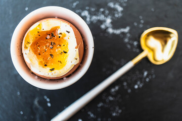Flat lay style food photo of a soft boiled egg in a white egg cup against a dark grey charcoal background.  Egg is seasoned with salt & pepper and a heart shaped teaspoon is arranged in the background