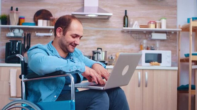 Disabled businessman in wheelchair during video call on laptop in kitchen. Wife cooking meal. Corporate man with paralysis handicap disability handicapped difficulties working after accident having