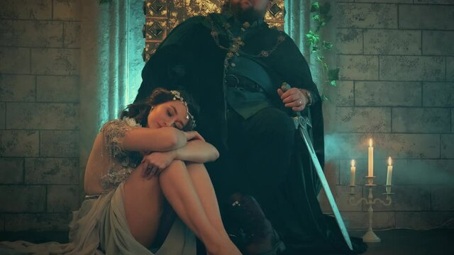 Themed wedding. beautiful woman queen sitting in feet medieval dictator king. Man on throne, holding sword. girl concubine slave of sultan. royal dress, vintage costume clothes gold crown. room castle