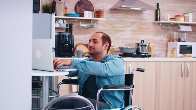 Handicapped businessman in wheelchair using laptop in kitchen. Corporate man with paralysis handicap disability handicapped difficulties working after accident having internet online video call
