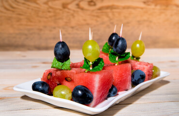 Canapes of watermelon, grapes, mint leaves, on a white plate, wooden table