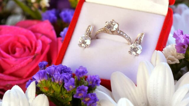 Close up view of gift present red box with a set of engagement ring and earrings on the background of a multi-colored beautiful bouquet of flowers. Marriage proposal, a new happy life
