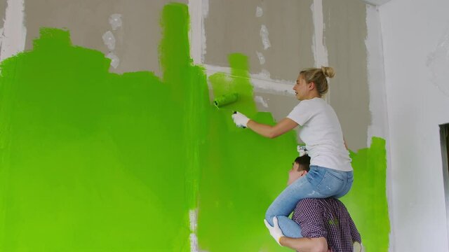 The girl sits on the shoulders of a strong guy and paints the wall with a roller in green. The guy Holds his girlfriend on his shoulders and shows her where to paint, the girl paints the drywall with