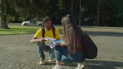 Handsome african male college student with dreadlocks helping pretty asian female classmate to pick up fallen textbooks at university campus, flirting while going together to high school lessons.