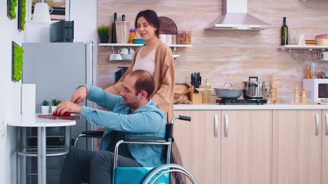 Disabled man in wheelchair chopping bell pepper in kitchen helping wife to cooking delicious meal. Guy with paralysis handicap disability handicapped difficulties helping in the kitchen