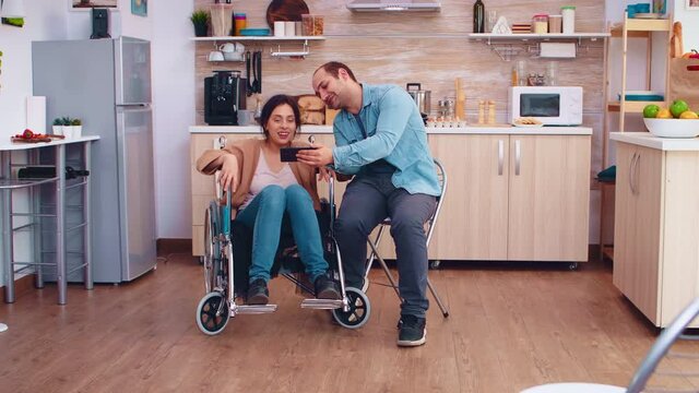 Cheerful disabled woman in wheelchair taking a selfie with husband in kitchen. Hopeful husband with handicapped disabled disability invalid paralysis handicap person next to him, helping her to