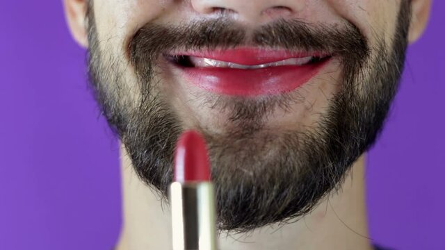 A young guy with a beard paints his lips with red lipstick. Close-up of a bearded man, he painted lips with bright lipstick. A bearded man puts red lipstick on his lips, smiles and smacks his lips.