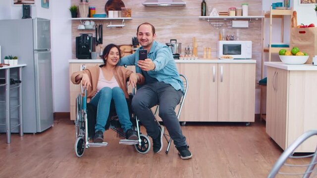 Positive woman in wheelchair and husband taking a selfie in kitchen using smartphone. Hopeful husband with handicapped disabled disability invalid paralysis handicap person next to him, helping her to