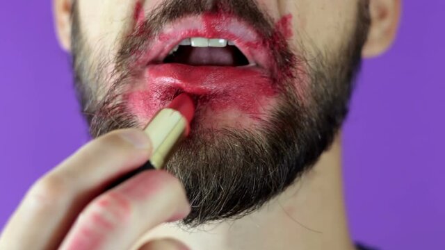 A young guy with a beard paints his lips with red lipstick. Close-up of a bearded man, he painted lips with bright lipstick. A bearded man puts red lipstick on his lips, smiles and smacks his lips.