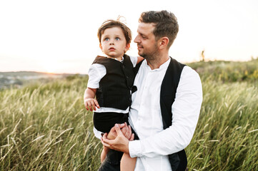 A little son in arms of his father with a scenic view on green hills on the background. Dad carries...