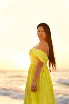 A girl in a long yellow dress walks by the sea in the early morning at dawn.