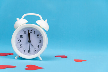 White alarm clock stands on a blue background with scattered red hearts, copy space