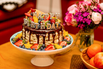 Delicious handmade layered cake decorated with Happy birthdays candles, chocolate glaze, strawberries, blueberries, carambola star fruit and ice cream cone on a table with flowers and fruits.