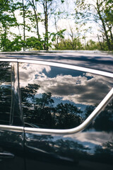 Reflection in window of car of the blue sky with clouds and forest trees in summer.