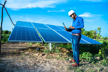 Young male engineer with laptop in hand standing near solar panels, agriculture farm land with...