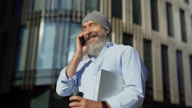 Panning medium shot of bearded senior man in hat walking down city street with laptop and takeaway coffee in his hand, talking on cell phone and laughing on sunny day