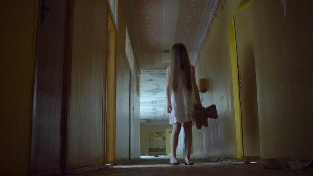 a girl with long black hair and a white dress is walking slowly in a dark corridor. The concept of a horror maniac. Halloween. she is holding a Teddy bear,