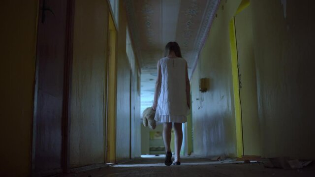 a girl with long black hair and a white dress is walking slowly in a dark corridor. The concept of a horror maniac. Halloween. she is holding a Teddy bear,