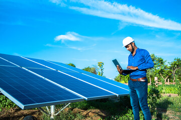 Young male engineer with laptop in hand standing near solar panels, agriculture farm land with clear blue sky background, Renewable energy, clean energy. - Powered by Adobe