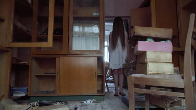 A demonic psycho child in a cursed abandoned house. A little girl dressed in a white dress with a bear toy looks scared at the 4K camera footage