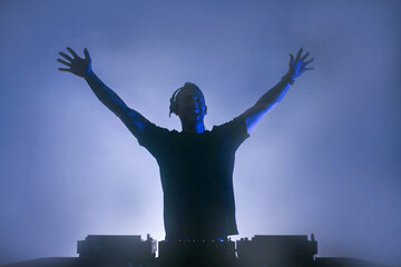 Black silhouette of a Male Disc jockey playing music raising two arms up.