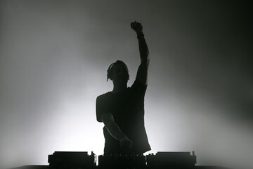 Disc jockey silhouette dressed in black playing music on a white wall in a smoky setting.