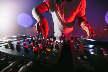 Close up of hands of a male DJ playing the mixer table for techno music in a dark colorful...
