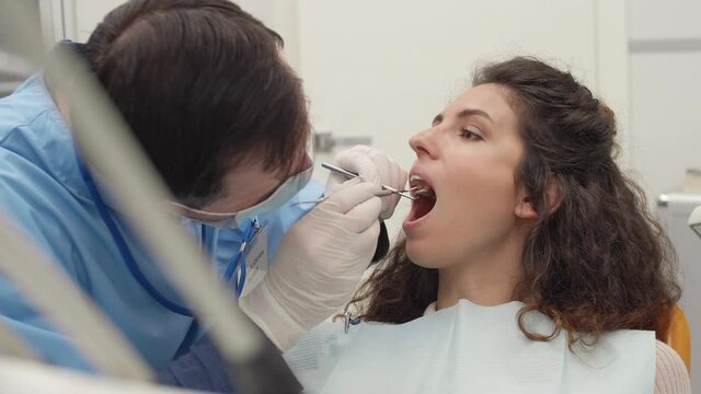 Close up of male dentist wearing medical overalls, mask and gloves holding special tools and checking teeth health of woman lying on dental chair
