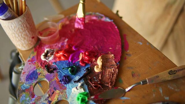 Female artist with thick paintbrush is mixing different coloured oil pink paints on palette. Close up view of female hand mixing colours. Colorful palette. High angle