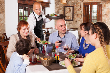 Cheerful waiter serving plates of salad to happy family having dinner at restaurant