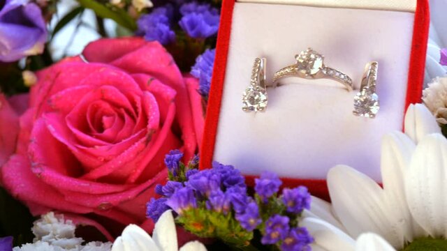 Close up view of gift present red box with a set of engagement ring and earrings on the background of a multi-colored beautiful bouquet of flowers. Marriage proposal, a new happy life