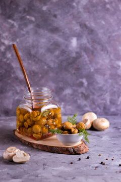Fermented canned champignon mushrooms in a white plate for appetizers and in a glass jar with garlic, bay leaf on a wooden stand