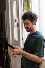 Young casual alternative model with moustache and curly hair using smartphone on a window indoors with natural light smiling facial expression and a corridor background with bike. 