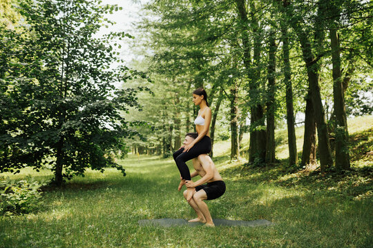 Happy couple engaged in sport in nature. Girl sitting on the shoulders of the guy, he doing squats. Outdoor workout open air. Man carrying woman on his shoulders and doing squats.