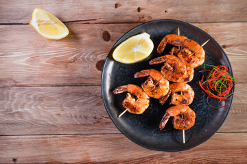 Appetizing fried shrimp on a skewer in a black plate on a wooden surface among lemon and seasonings. The concept of fast and wholesome food