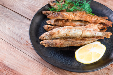 Appetizing fried capelin on a black plate stands on a wooden surface. Simple rustic food concept