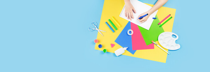 Hands drawing over color paper, scissors, pencils paint and various school stationery on blue yellow background. Flat lay with copy space for back to school or education and craft concept banner