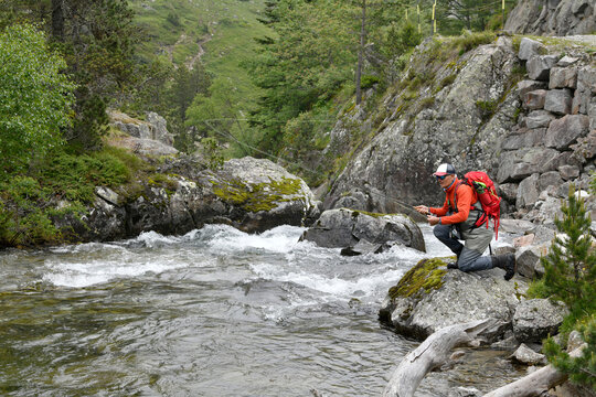 Fly Fisherman Trout Fishing With A Hiking Backpack And An Orange Jacket In The High Mountains In Summer