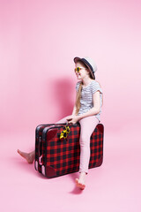 Travel: A little girl in a straw hat and a medical mask is sitting on a big red suitcase and waiting for travel.