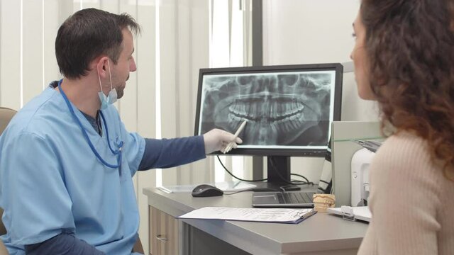 Medium shot of Caucasian woman and middle-aged male dentist sitting in dental office and discussing x-ray image of oral cavity on the screen