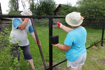 An elderly russian farmers family couple builds an outdoor fence, a woman paints the base with anti corrosion black paint, a man tampers the posts with a large tamp bar  on Sunny summer day