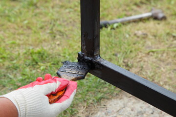 Anticorrosive painting of welded joints metal constructions, a hand in a work glove paints the welded fence frame with black bituminous paint closeup on green grass and blurred hammer background