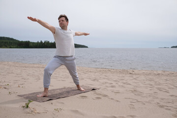 Caucasian young man doing yoga exercises on beach in summer time. Warrior pose or virabhadrasana