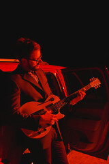 Businessman in a suit playing an electric guitar at night in the red light near the car