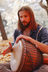 Young hippie man playing on a djembe drum in autumn in the forest.