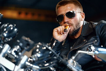 Cool bearded man biker in sunglasses sitting on his motorbike and smoking cigar