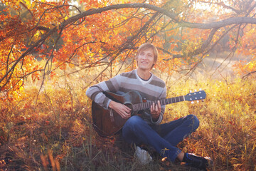 Young caucasian man in a sweater playing an acoustic guitar in the autumn forest.