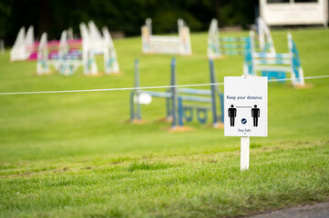 A Covid-19 social distancing sign staked into grass with an equestrian show jumping outdoor event in the background