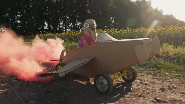 Little happy pilot girl 2 years old in a pilot hat sits in a cardboard plane, dad pulls a cart with a cardboard airplane on wheels along a country road